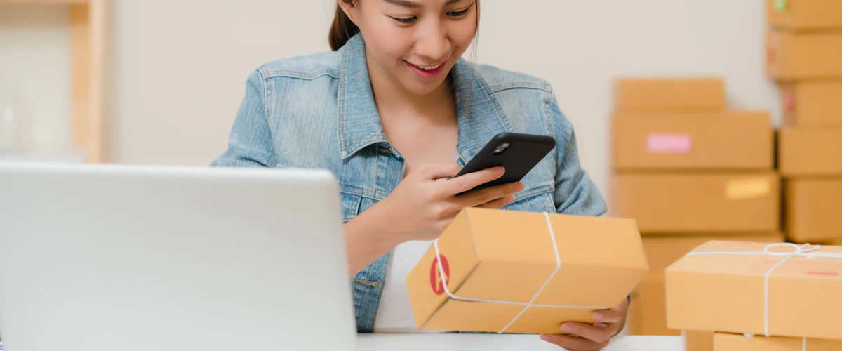 Emprendedora revisa paquetes con un móvil y portátil en su pequeño negocio