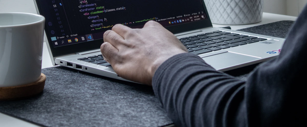 Persona programando en un portátil situado junto a una planta y una taza